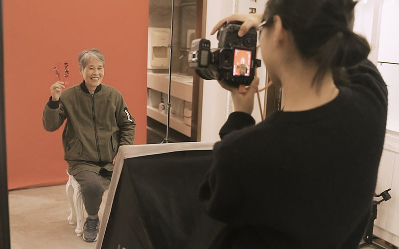 Person being photographed in a studio while holding a red envelope against a red backdrop.